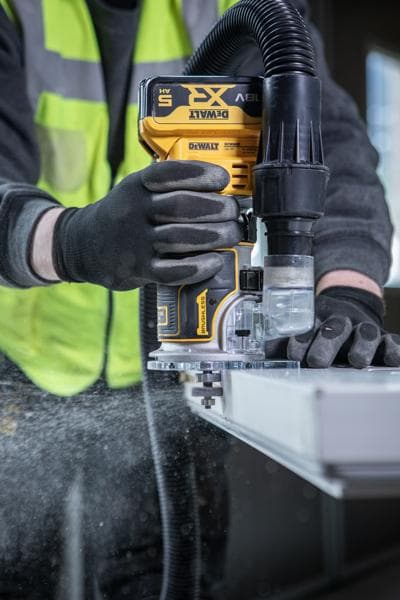 A person wearing safety gloves and a high-visibility vest uses a DEWALT cordless router tool with dust extraction, working on the edge of a piece of wood or another flat material.