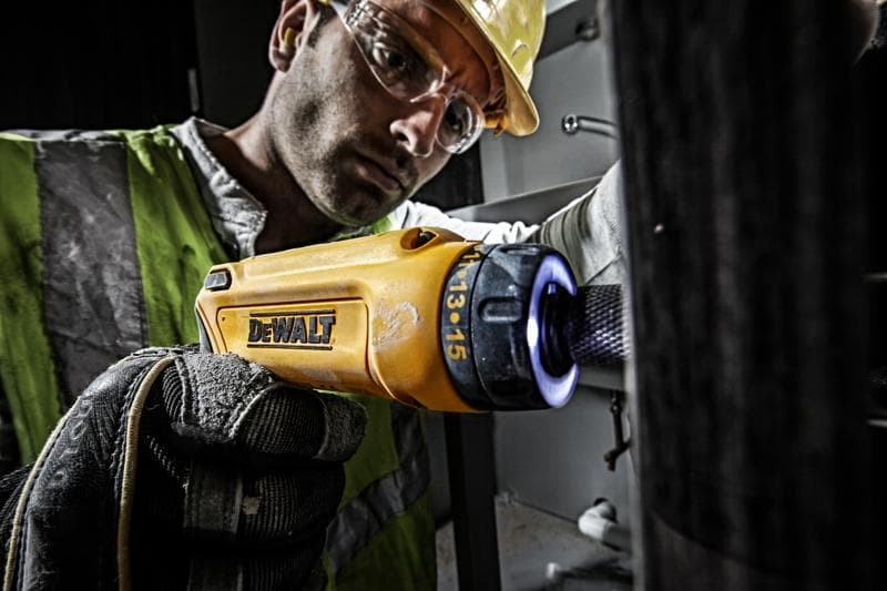 A person wearing a reflective safety vest and gloves uses a yellow DEWALT cordless screwdriver (model DCF680G2) for maintenance work indoors. The focus is on the tool and the user's gloved hand. The person's face is blurred for privacy.