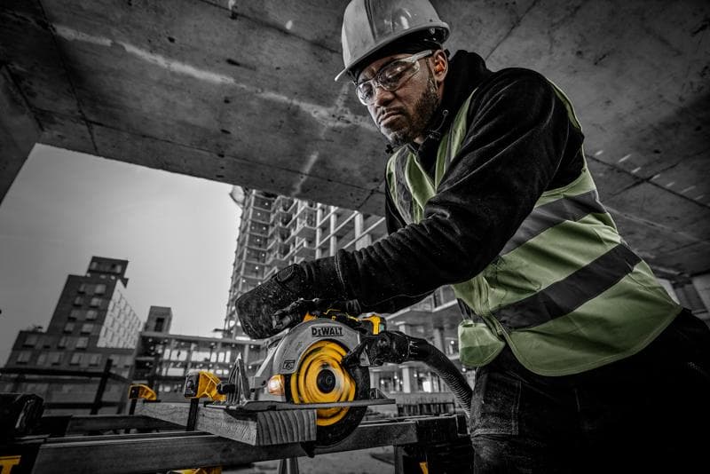 A construction worker wearing a safety vest and helmet operates a DEWALT circular saw (model DCS565NT) to cut a wooden plank at a building site. The background shows concrete structures and high-rise buildings under construction.