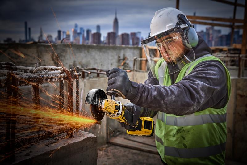 A person wearing protective gear, including a helmet, face shield, gloves, and a high-visibility vest, is using a yellow DEWALT cordless angle grinder (SKU: DCG405P2) to cut through metal rebar on a construction site. Sparks are flying from the cutting area. The city skyline is visible in the background.