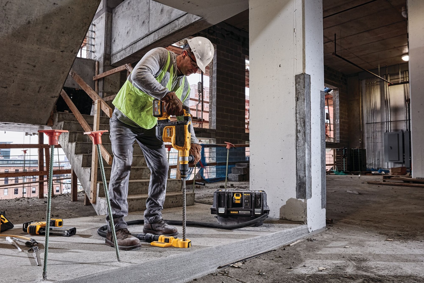 A worker in a construction site wearing a safety vest and helmet is using a DEWALT cordless power tool to drill into a concrete surface. The DEWALT DCV586MT2 dust extractor is visible on the floor beside the worker.
